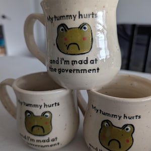 May include: Three beige ceramic mugs with a frog illustration and the text "My tummy hurts and I'm mad at the government." The frog is green with a sad expression. The mugs have a speckled appearance and a curved handle.