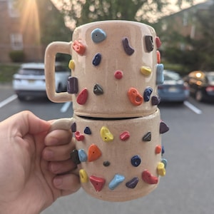 May include: Two beige ceramic mugs stacked, each decorated with colourful, textured climbing holds. The mugs have a speckled finish and a handle. The climbing holds are in various shapes and colours, including red, blue, yellow, and orange.