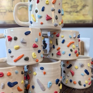 Op de afbeelding: Een stapel crèmekleurige keramische mokken met kleurrijke, getextureerde klimgrepen. De grepen hebben verschillende vormen en kleuren, waaronder rood, blauw, geel en oranje. De mokken hebben een eenvoudig, afgerond ontwerp met een oor.