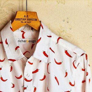 May include: White shirt with a red chilli pepper pattern. The shirt has the text "richardesign" on the label and is hanging on a wooden hanger. The hanger has the text "A.B. CHRISTIAN JOHNSSON TEL. 48 05BY".