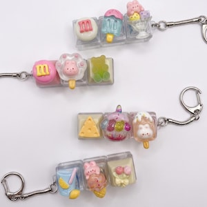 May include: A collection of colorful, miniature food-themed keychains. Each keychain features a series of small, clear blocks containing various food items like ice cream, popsicles, and a slice of cheese. The keychains have silver-colored metal hardware.
