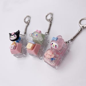 May include: Three keychains with cartoon character figures. Each keychain features a clear, rectangular block with a pink base. The characters include a black and white figure, a light green figure, and a pink figure. Each keychain has a silver clasp.