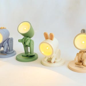 May include: Four different colored desk lamps shaped like animals. The lamps are blue, green, white, and beige. The blue lamp is shaped like a robot, the green lamp is shaped like a robot, the white lamp is shaped like a rabbit, and the beige lamp is shaped like a rabbit. The lamps have a light on inside that is turned on.