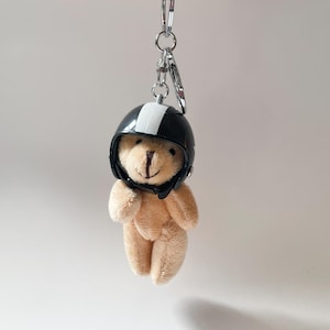 May include: A small, tan teddy bear wearing a black and white helmet. The bear is attached to a silver key chain.
