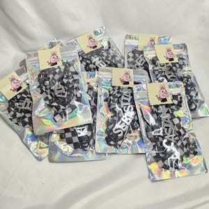 May include: Multiple clear, iridescent pouches filled with black and white chequered accessories. Each pouch has a small, square tag featuring a cartoon pig. Some accessories have names such as "Sebastian" and "Castasia" in a silver font.