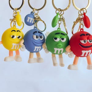 Puede incluir: Cuatro llaveros coloridos con personajes de dibujos animados de caramelos. Cada llavero tiene un color diferente: amarillo, azul, verde y rojo. Los personajes tienen brazos, piernas y pies blancos, y la letra "M" en el pecho.