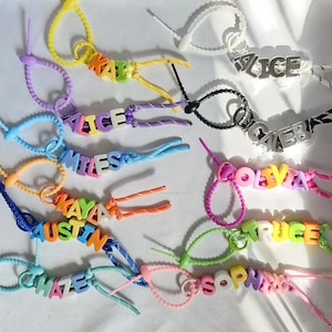 May include: A collection of personalized zipper pulls featuring colorful plastic letter beads spelling names like Kai, Alice, Miles, Kaya, Austin, Haze, Caleb, Olivia, Truce, and Sophia, attached to various patterned and solid colored nylon cords.