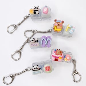 May include: Several novelty keychains with silver-tone hardware. Each keychain features a clear plastic block with colorful miniature figures, including pandas, a bear with a duck floatie, and flip-flops. Some blocks have the words "Lucky" and "Happy".