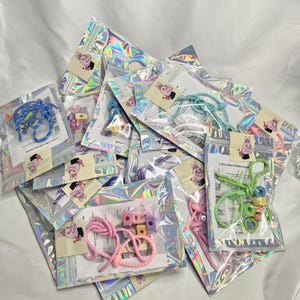 Könnte beinhalten: Eine Sammlung kleiner, klarer, irisierender Plastiktüten, gefüllt mit bunten Bastelmaterialien. Jede Tüte hat einen Aufkleber mit einem Cartoon-Schwein mit Hut. Die Tüten enthalten Artikel wie Seile, Perlen und Anleitungen zur Schmuckherstellung.