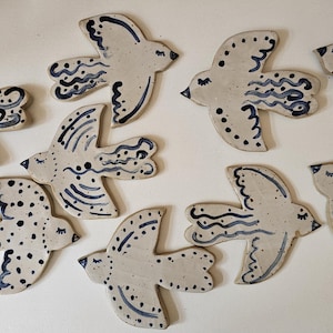 Könnte beinhalten: Eine Sammlung von neun flachen, vogelgeformten Keramikornamenten in hellem Beige. Jeder Vogel weist einzigartige, handgemalte Details in Dunkelblau auf, darunter Punkte, wellenförmige Linien und geschwungene Muster auf den Flügeln und Körpern.