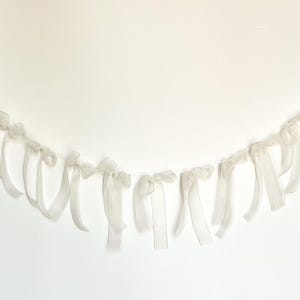 May include: A garland of white organza ribbon bows hangs against a white background. The bows are evenly spaced along a thin string, creating a delicate and airy decorative element. The ribbon has a slight sheen.