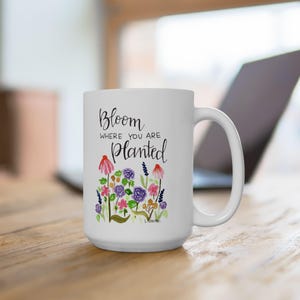 Pode incluir: Caneca de cerâmica branca com um design floral e o texto "Bloom where you are planted". As flores em estilo aquarela incluem flores rosa, roxas e azuis. A caneca tem uma alça grande e está em uma superfície de madeira.
