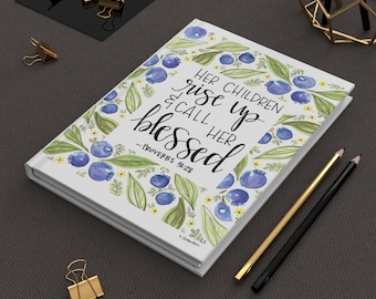 May include: A white journal with a watercolour design of blue berries and green leaves. The text on the cover reads "Her children rise up and call her blessed - Proverbs 31:28".