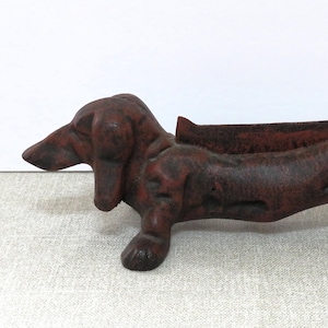 May include: A dark brown, cast iron dachshund-shaped boot scraper with a textured, weathered finish. The dog figure is depicted in a standing position with a long body, floppy ears, and a curled tail, featuring a flat, brush-like insert along its back.