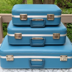 May include: Three stacked vintage blue suitcases with white trim and gold-colored latches and handles. The suitcases are arranged from largest to smallest, creating a visually appealing display. The background features a natural outdoor setting.
