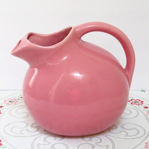 May include: A solid pink ceramic pitcher with a rounded, spherical body, a curved handle on the right side, and a wide, molded pouring spout. The surface of the pitcher has a smooth, glossy finish that reflects light, highlighting its curved shape.