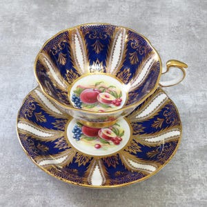 May include: A vintage teacup and saucer set. The set features a deep blue and gold design with white accents. The cup and saucer have a scalloped edge and a fruit illustration in the center. The handle of the cup is gold.