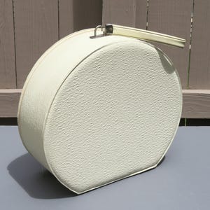 May include: Off-white, round vintage hatbox with a textured surface. The hatbox has a matching handle and a metal clasp. The hatbox is sitting on a gray surface.
