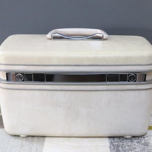 May include: Cream-colored vintage vanity case with a silver handle and trim. The rectangular case has a textured surface, a silver lock, and small feet. A classic travel accessory.