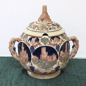 May include: A ceramic tureen with a lid, featuring a detailed design of castles and grapevines. The tureen has a beige base with brown and blue accents. The handles are intricately sculpted. The lid has a small tower-like structure on top.
