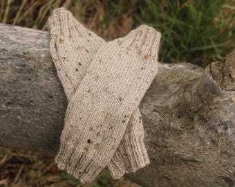 May include: A pair of beige knitted fingerless gloves with a speckled pattern. The gloves are laid out on a piece of weathered wood.