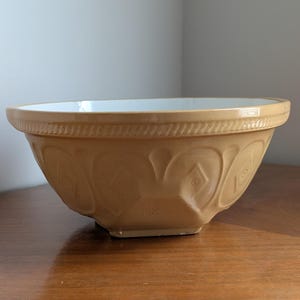May include: A tan ceramic mixing bowl with a white interior. The exterior features a textured rope pattern around the rim and a repeating embossed diamond and arch design along the sides. The bowl has a wide, circular opening and a sturdy base.