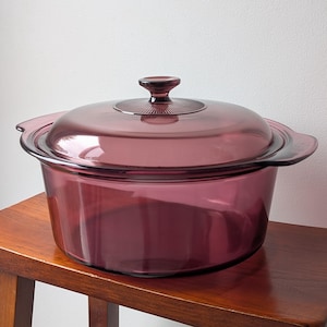 May include: A round, translucent purple glass casserole dish with integrated side handles and a matching glass lid. The lid features a central knob and a subtle, radiating sunburst pattern around the handle, showcasing a smooth, polished finish.