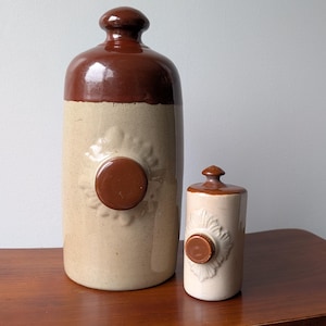 May include: Two cylindrical stoneware hot water bottles with a glossy brown glaze on the top and neck, and a light beige body. Each bottle features a central circular brown screw cap surrounded by a moulded sunburst or floral relief pattern.