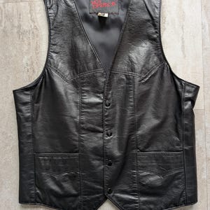 Może przedstawiać: Czarna skórzana kamizelka z dekoltem w serek, dwiema przednimi kieszeniami i zapięciem na guziki. Kamizelka ma teksturowany wygląd i satynową podszewkę. Etykieta wewnątrz kamizelki głosi "The Leather Shop".