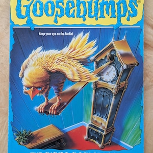 May include: A paperback book titled "Goosebumps: The Cuckoo Clock of Doom" by R.L. Stine. The cover features a menacing bird perched on a wooden plank, a grandfather clock, and the text "Keep your eye on the birdie!" in a blue and yellow design.