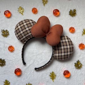 May include: A headband featuring two round ears covered in a brown, tan, and black plaid fabric. Centered between the ears is a large, ribbed bow in a solid burnt orange color. The headband frame is wrapped in a thin black material.