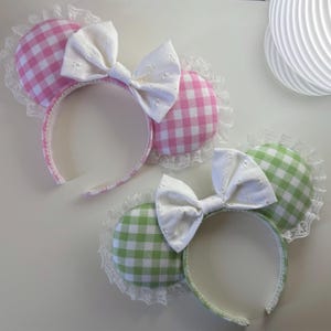 May include: Two headbands featuring circular ears with a pink and white gingham pattern and a green and white gingham pattern respectively. Each headband is adorned with a white eyelet fabric bow and delicate white lace trim along the edges.