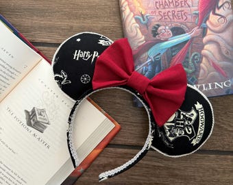 May include: Black and white Harry Potter themed mouse ears with a red bow. The ears feature the Hogwarts crest and the words "Harry Potter" and "Hogwarts".