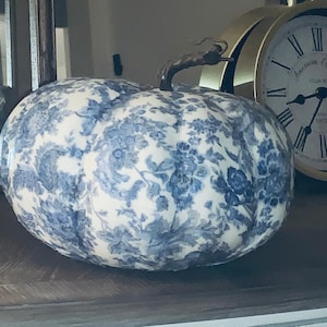 May include: A white pumpkin with a blue and white floral pattern. The pumpkin has a stem and is sitting on a wooden surface.