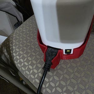 May include: A white cylindrical container sits inside a red 3D-printed plastic base. A black power cord is plugged into the side of the red base, and a small rectangular power switch with a glowing yellow light is visible next to the cord connection.