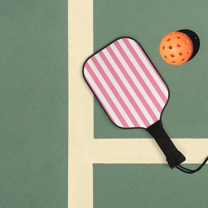 Könnte beinhalten: Ein rosa-weiß gestreiftes Pickleball-Schläger mit schwarzem Griff und schwarzem Band. Der Schläger liegt auf einem grünen Pickleball-Platz mit einer weißen Linie.