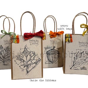 May include: Four brown kraft paper gift bags featuring black ink botanical sketches of an elm leaf, a maple leaf, an acorn, and an oak leaf. Each bag is printed with the text Thanksgiving Blessings and includes a colorful crinkled ribbon tie.