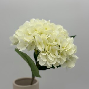 May include: A single stem of artificial hydrangea flowers in a beige vase. The flower head is a cluster of creamy white petals, with a green stem and leaf. The vase is a simple, modern design.