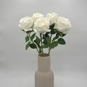 Può includere: Un bouquet di sei rose artificiali color crema con foglie e steli verdi, disposto in un vaso cilindrico alto in ceramica beige con superficie a coste. I fiori presentano petali stratificati e sono posizionati in verticale in una composizione.