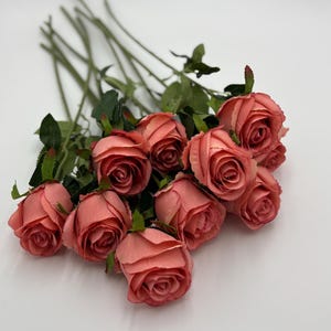 May include: A bouquet of artificial roses in a muted coral-pink shade. Each flower features layered, soft-edged petals and is attached to a long, slender green stem with small, pointed green leaves extending from the base of the bloom.