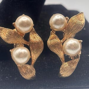 May include: A pair of gold tone clip-on earrings with three faux pearls on each earring. The earrings are shaped like leaves.