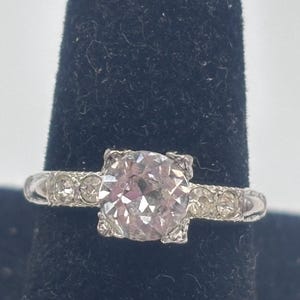 May include: A silver ring with a large, round, light pink gemstone at the center. Smaller clear gemstones are set along the band on either side of the center stone. The ring is displayed on a black surface.