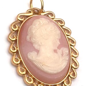 May include: Oval pendant with a cameo of a woman's profile in cream against a pink background. The cameo is set in a gold-colored, ornate frame with a loop at the top for a chain. The frame has a decorative, swirling design.