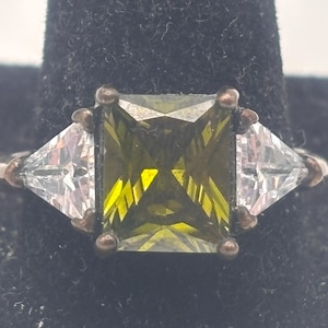 May include: A gold ring with a large, square, green gemstone surrounded by two smaller, triangular, clear gemstones.