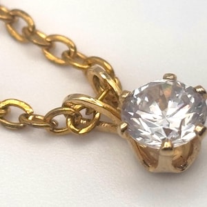 May include: A gold-colored necklace with a large, clear, round gemstone pendant. The pendant is set in a gold-colored prong setting. The chain is a delicate gold-colored link chain.