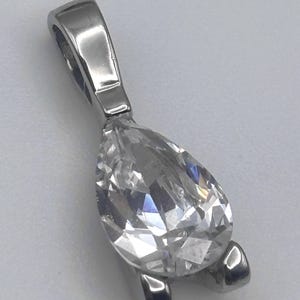May include: A pear-shaped, clear crystal pendant with a polished silver-tone bail and setting. The crystal is faceted, reflecting light. The pendant is designed to be worn as a necklace.