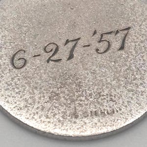 May include: Close-up of a silver-toned, circular pendant with a textured surface. The inscription "6-27-'57" is etched in a cursive font. The word "STERLING" is also visible.