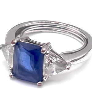 May include: A silver ring featuring a large, rectangular, deep blue gemstone flanked by two smaller, triangular, clear gemstones. The ring band has a multi-strand design, creating a unique and elegant look. A piece of jewelry.