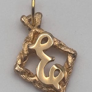 May include: A gold-colored pendant featuring the letter "e" in a cursive font, set within a textured, diamond-shaped frame. The pendant includes a small loop for a chain. The design has a vintage aesthetic.