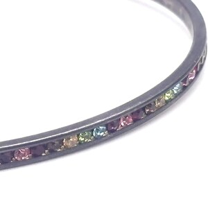 May include: A thin, dark gray metal bangle bracelet featuring a channel setting that holds a continuous row of small, round, multi-colored gemstones in shades of pink, purple, blue, green, and yellow, set flush within the band's surface.